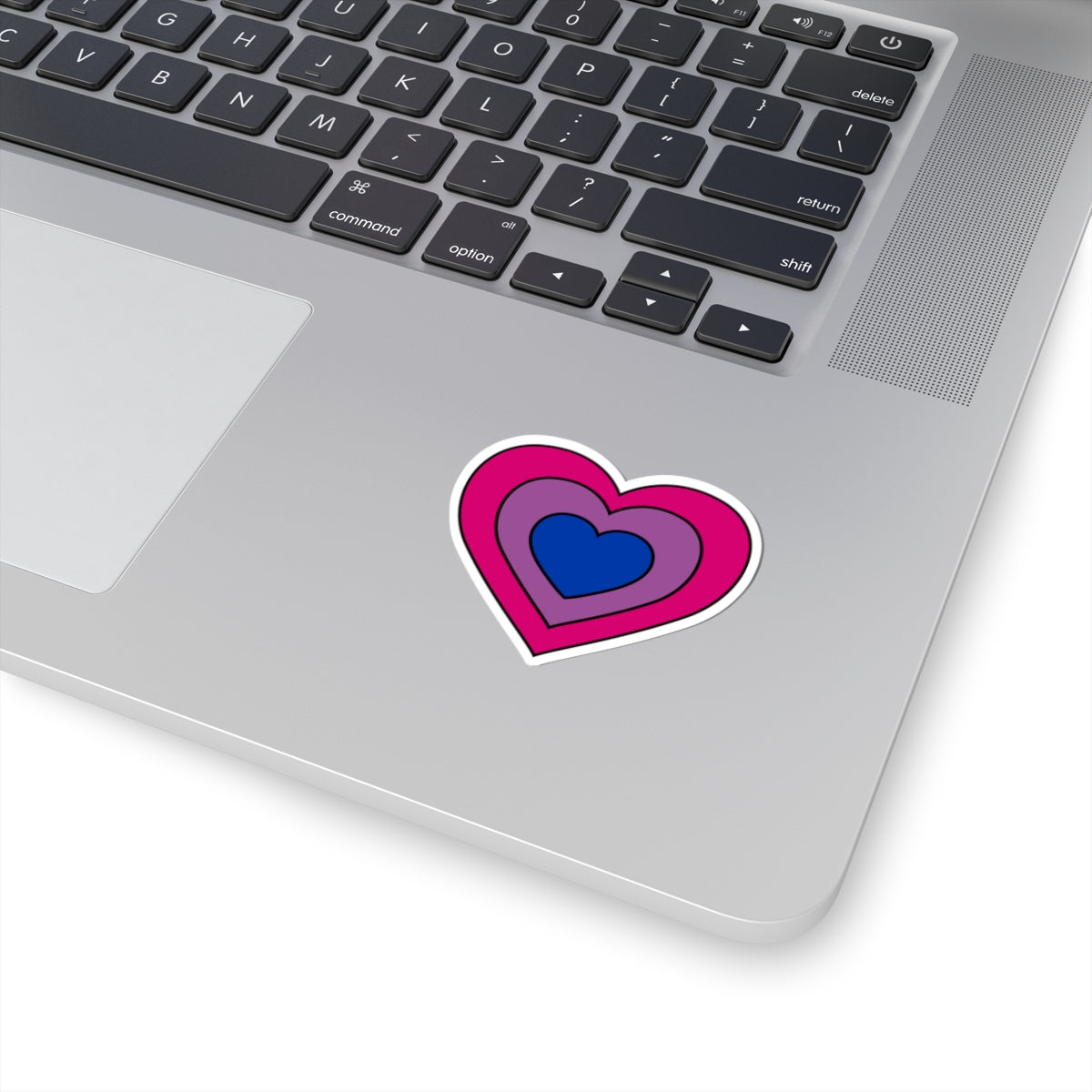 Close-up of the bottom right corner of a silver laptop featuring a 2 inch by 2 inch pride flag heart sticker in a Bisexual flag style applied to the surface.