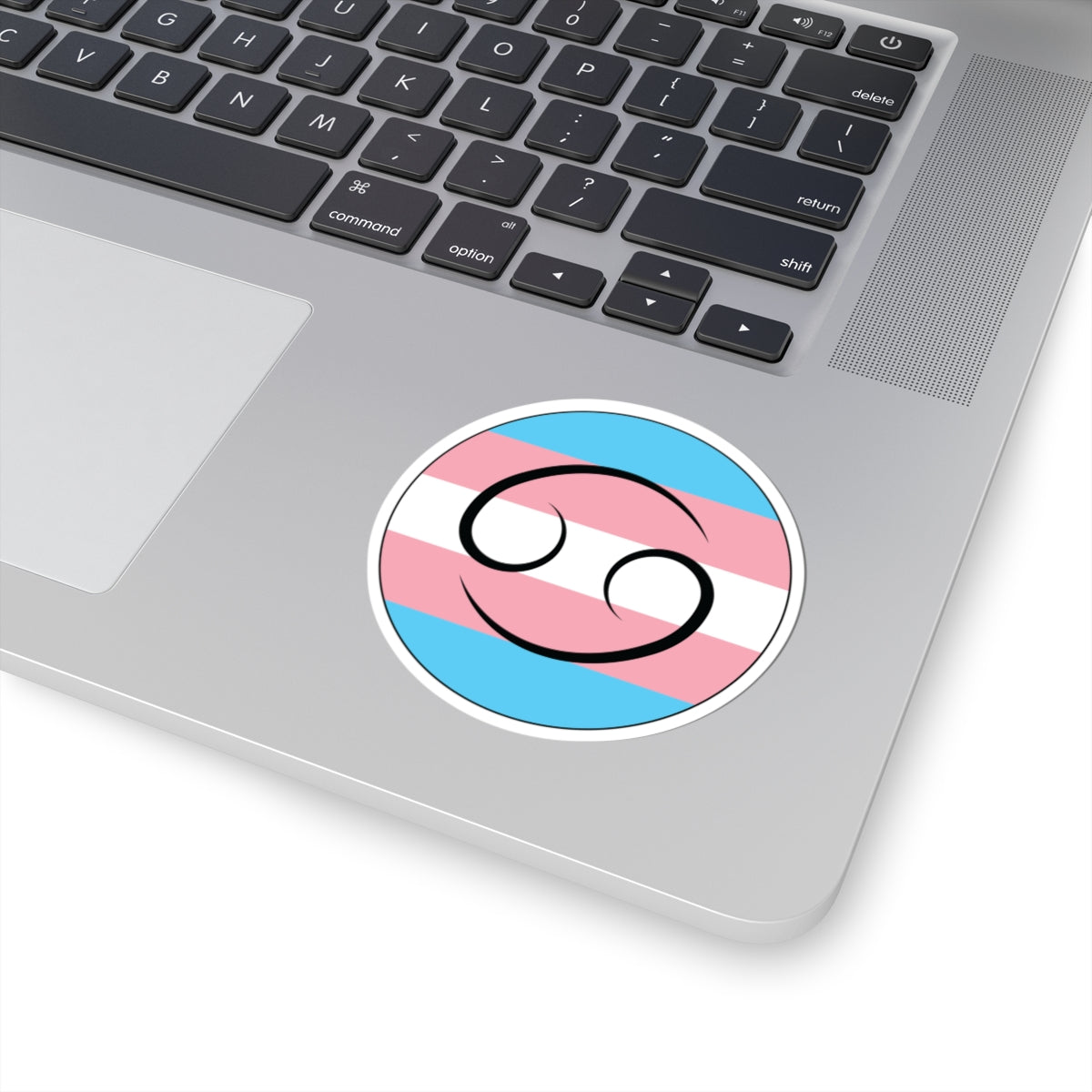 Close-up of the bottom right corner of a silver laptop featuring a 3 inch by 3 inch circular zodiac sticker with a centered Cancer glyph on a Transgender pride flag style background applied to the surface.