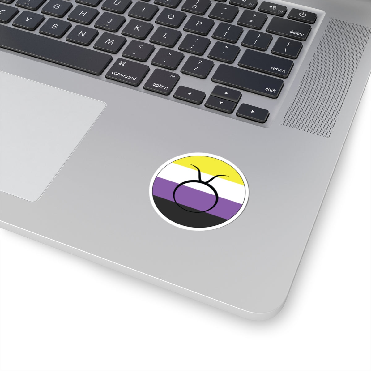 Close-up of the bottom right corner of a silver laptop featuring a 2 inch by 2 inch circular zodiac sticker with a centered Taurus glyph on a Nonbinary pride flag style background applied to the surface.