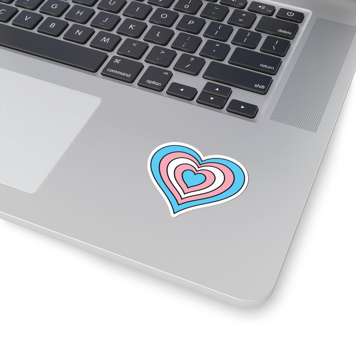Close-up of the bottom right corner of a silver laptop featuring a 2 inch by 2 inch pride flag heart sticker in a Transgender flag style applied to the surface.