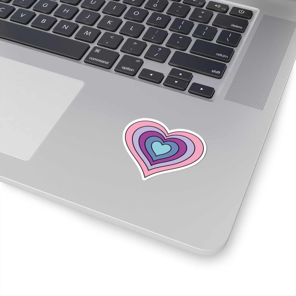 Close-up of the bottom right corner of a silver laptop featuring a 2 inch by 2 inch pride flag heart sticker in an Omnigender flag style applied to the surface.