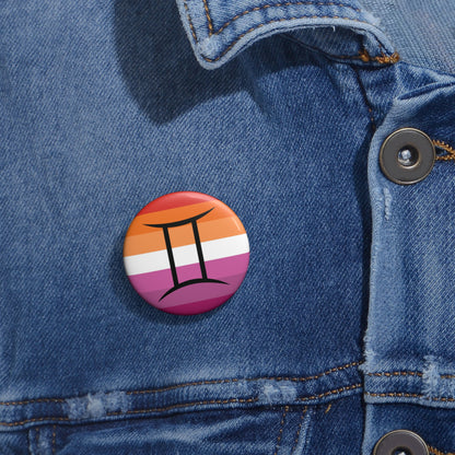 Close-up of a white circular pride button featuring the Gemini zodiac glyph centered over a lesbian pride flag background in oranges, white, and pinks attached to a blue denim jacket at the right chest with the button clearly visible.