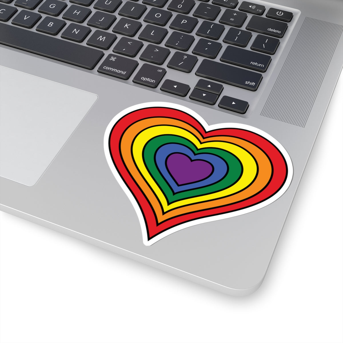 Close-up of the bottom right corner of a silver laptop featuring a 4 inch by 4 inch pride flag heart sticker in a rainbow pride flag style applied to the surface.