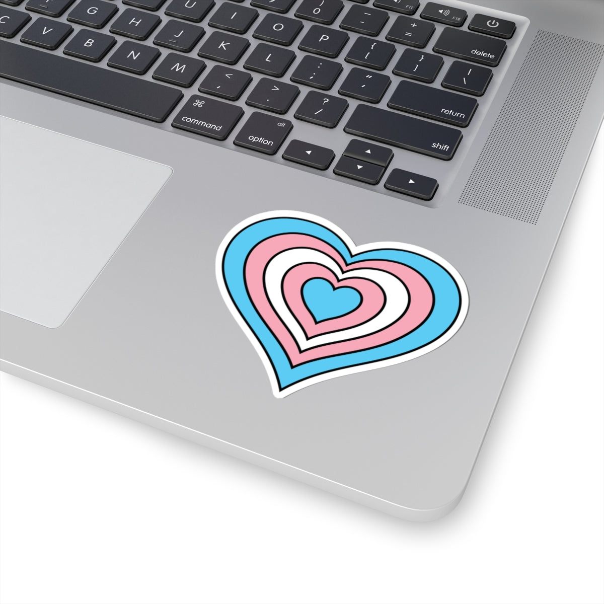 Close-up of the bottom right corner of a silver laptop featuring a 3 inch by 3 inch pride flag heart sticker in a Transgender flag style applied to the surface.