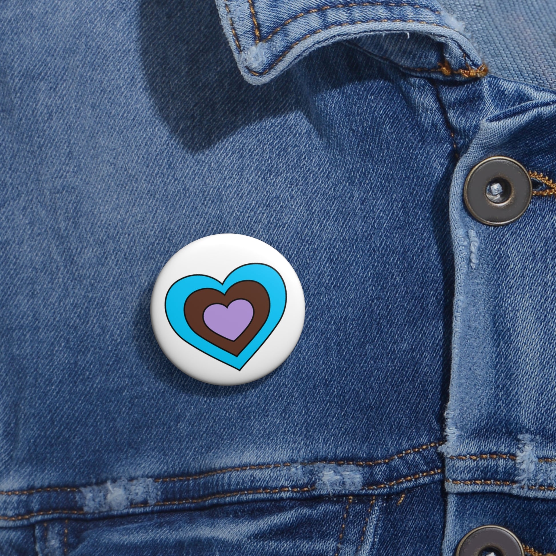 Close-up of a white circular pride button featuring a nested heart design inspired by the Androsexual pride flag in blue, brown and lavender, attached to a blue denim jacket at the right chest with the button clearly visible.