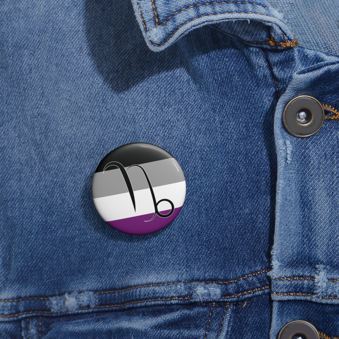 Close-up of a white circular pride button featuring the Capricorn zodiac glyph centered over an asexual pride flag background in black, gray, white, and purple, attached to a blue denim jacket at the right chest with the button clearly visible.