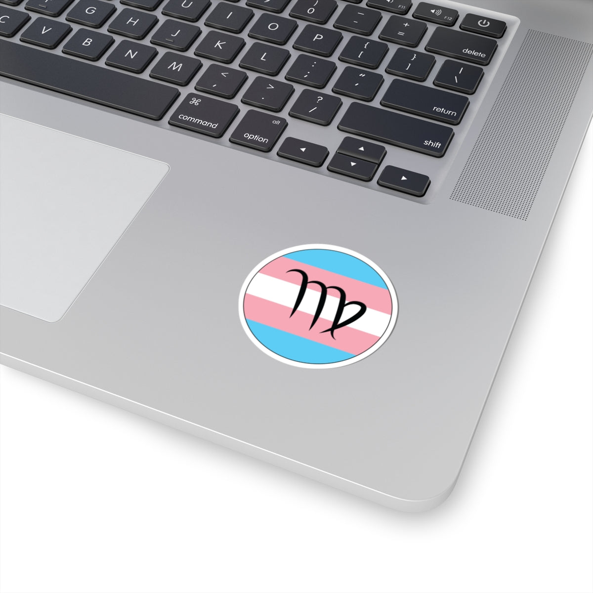 Close-up of the bottom right corner of a silver laptop featuring a 2 inch by 2 inch circular zodiac sticker with a centered Virgo glyph on a Transgender pride flag style background applied to the surface.