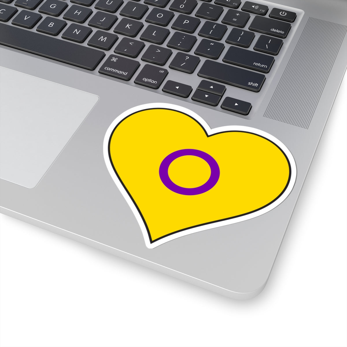 Close-up of the bottom right corner of a silver laptop featuring a 4 inch by 4 inch pride flag heart sticker in an Intersex flag style applied to the surface.