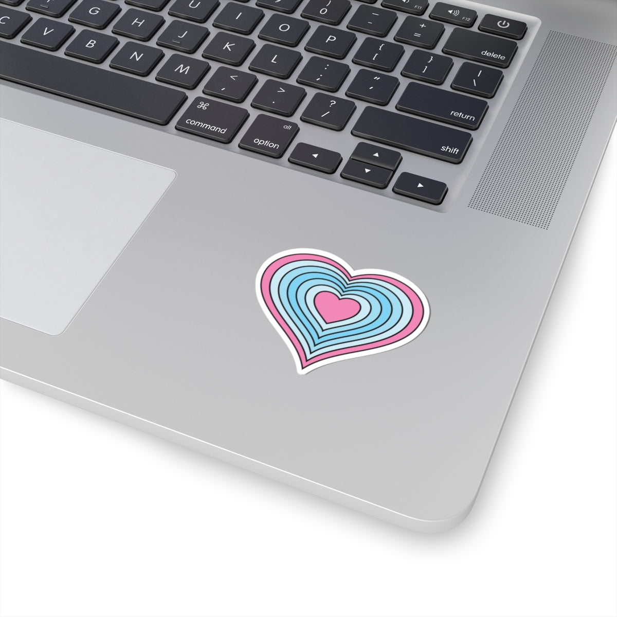 Close-up of the bottom right corner of a silver laptop featuring a 2 inch by 2 inch pride flag heart sticker in a Transmasc flag style applied to the surface.