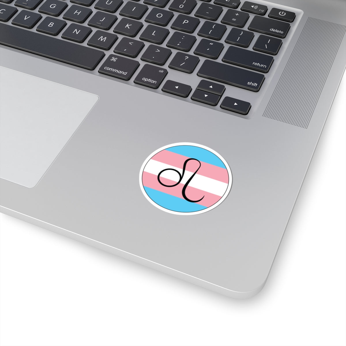 Close-up of the bottom right corner of a silver laptop featuring a 2 inch by 2 inch circular zodiac sticker with a centered Leo glyph on a Transgender pride flag style background applied to the surface.