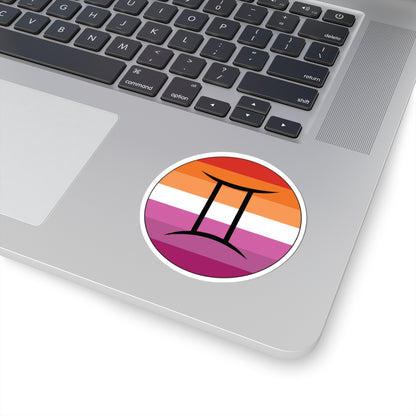 Close-up of the bottom right corner of a silver laptop featuring a 3 inch by 3 inch circular zodiac sticker with a centered Gemini glyph on a Lesbian pride flag style background applied to the surface.