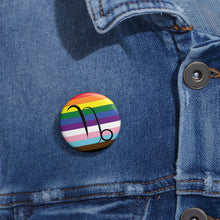 Close-up of a white circular pride button featuring the Capricorn zodiac glyph centered over a Progress Pride flag background in rainbow stripes with black, brown, light blue, pink, and white elements, attached to a blue denim jacket at the right chest with the button clearly visible.