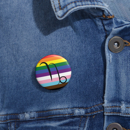 Close-up of a white circular pride button featuring the Capricorn zodiac glyph centered over a Progress Pride flag background in rainbow stripes with black, brown, light blue, pink, and white elements, attached to a blue denim jacket at the right chest with the button clearly visible.