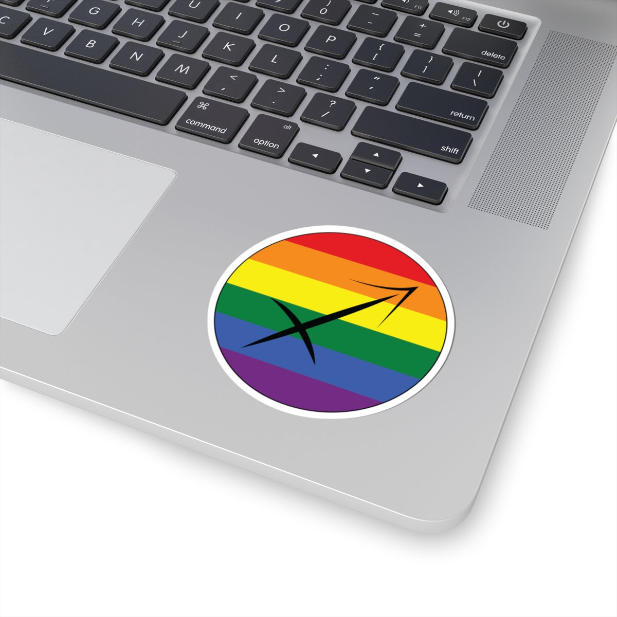 Close-up of the bottom right corner of a silver laptop featuring a 3 inch by 3 inch circular zodiac sticker with a centered Sagittarius glyph on a Rainbow pride flag style background applied to the surface.