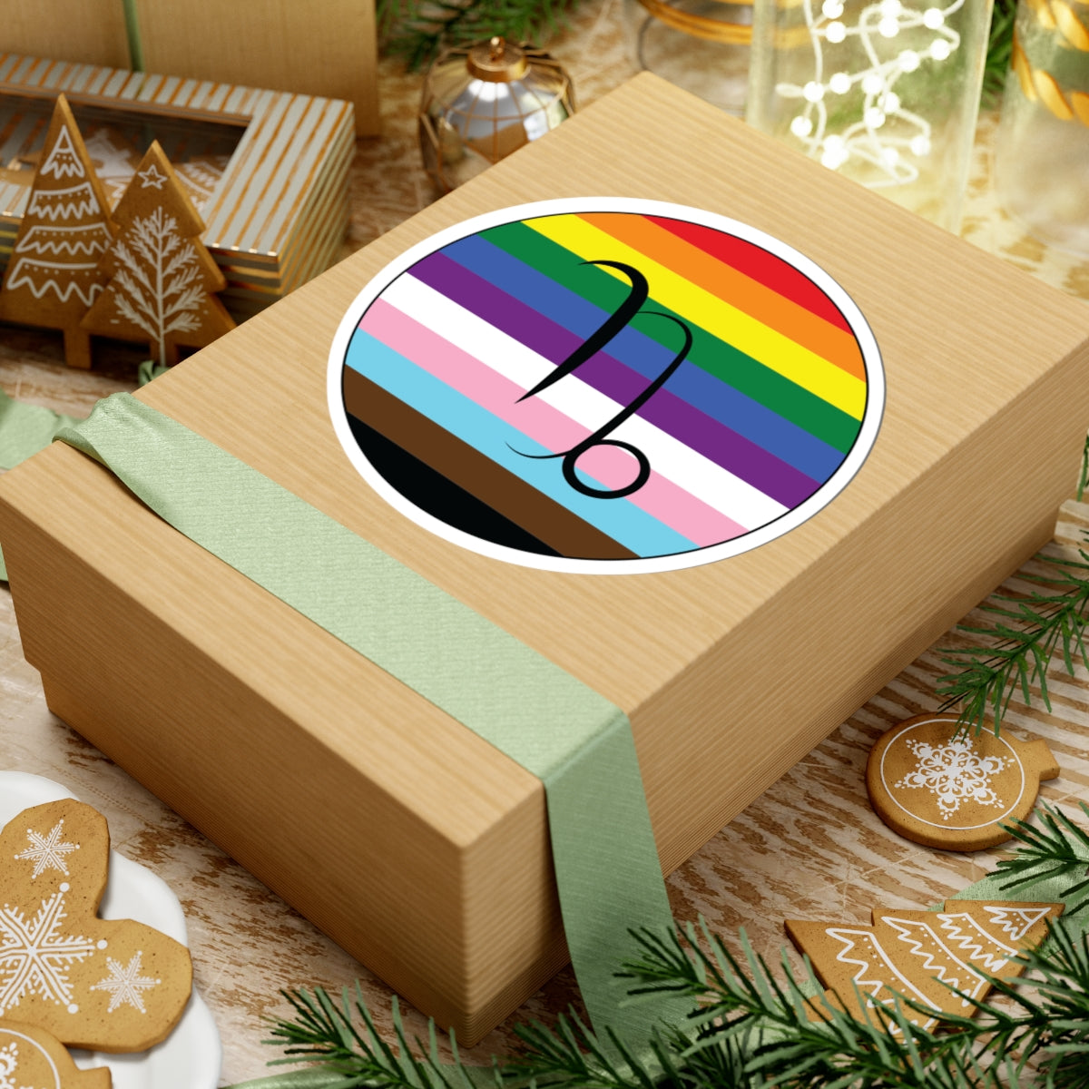 Gift box with a green ribbon on a festive background with cookies and Christmas decorations. Capricorn Progress pride sticker centered on the box.
