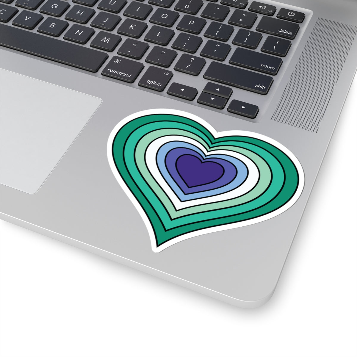 Close-up of the bottom right corner of a silver laptop featuring a 4 inch by 4 inch pride flag heart sticker in a Gay Men flag style applied to the surface.