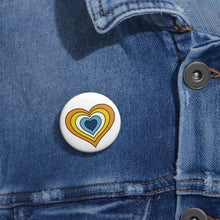 Close-up of a white circular pride button featuring a nested heart design inspired by the Aroace pride flag in yellows, white and blues, attached to a blue denim jacket at the right chest with the button clearly visible.