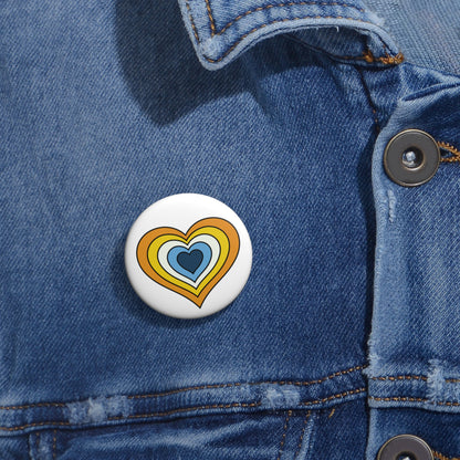 Close-up of a white circular pride button featuring a nested heart design inspired by the Aroace pride flag in yellows, white and blues, attached to a blue denim jacket at the right chest with the button clearly visible.