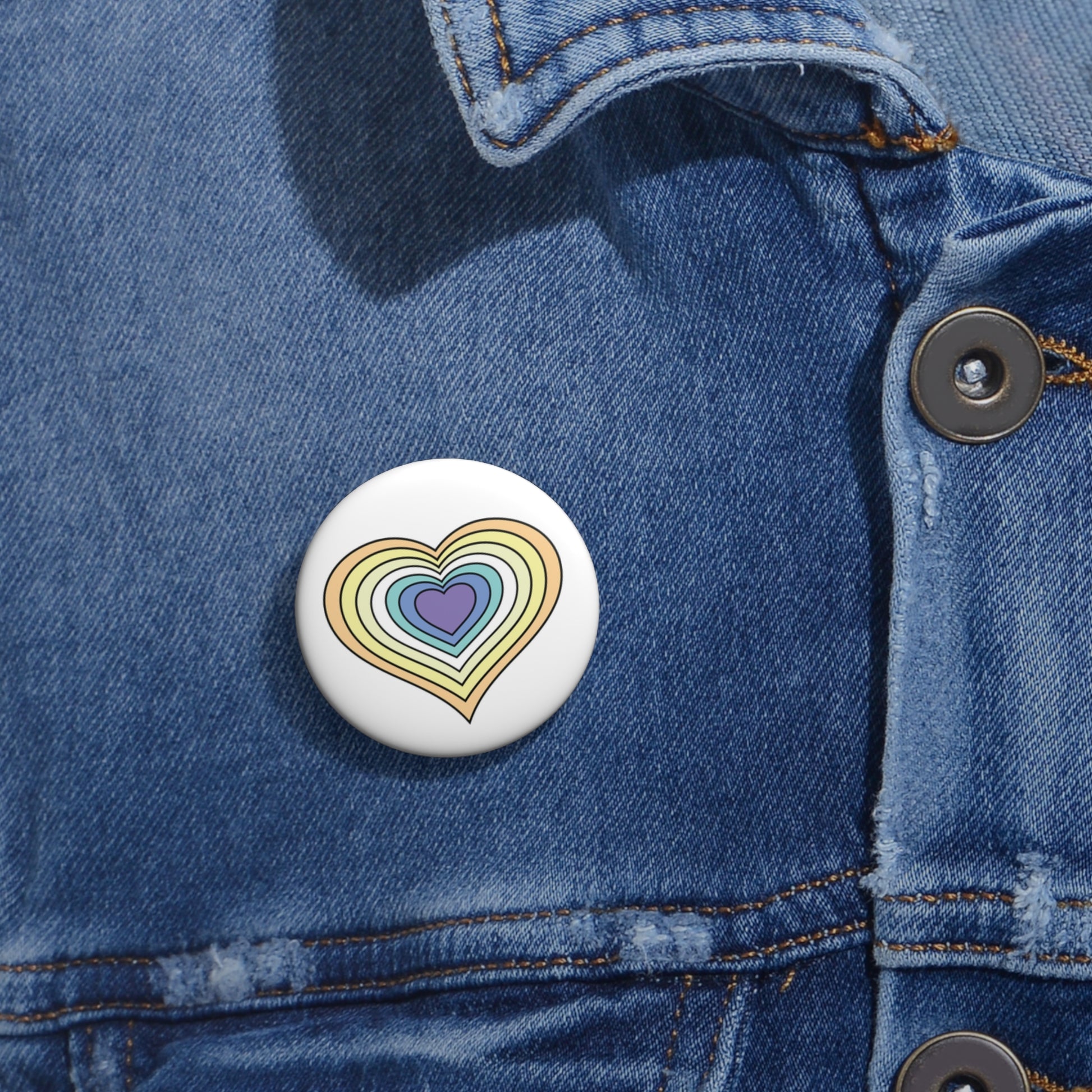 Close-up of a white circular pride button featuring a nested heart design inspired by the Genderfaun pride flag in yellows, white and blues, attached to a blue denim jacket at the right chest with the button clearly visible.