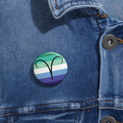 Close-up of a white circular pride button featuring the Aires zodiac glyph centered over a gay men (MLM) pride flag background in greens/teals, white, and blues, attached to a blue denim jacket at the right chest with the button clearly visible.
