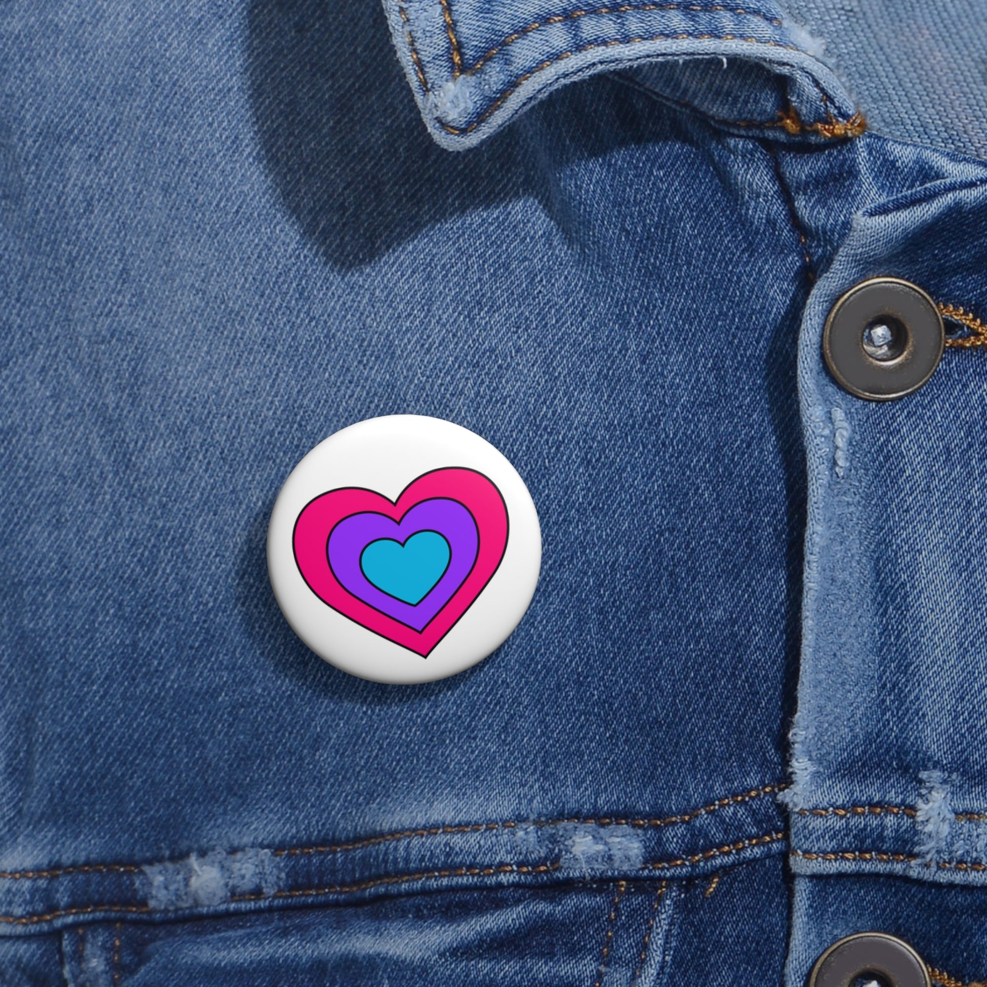 Close-up of a white circular pride button featuring a nested heart design inspired by the Androgyne pride flag in pink, purple and blue, attached to a blue denim jacket at the right chest with the button clearly visible.
