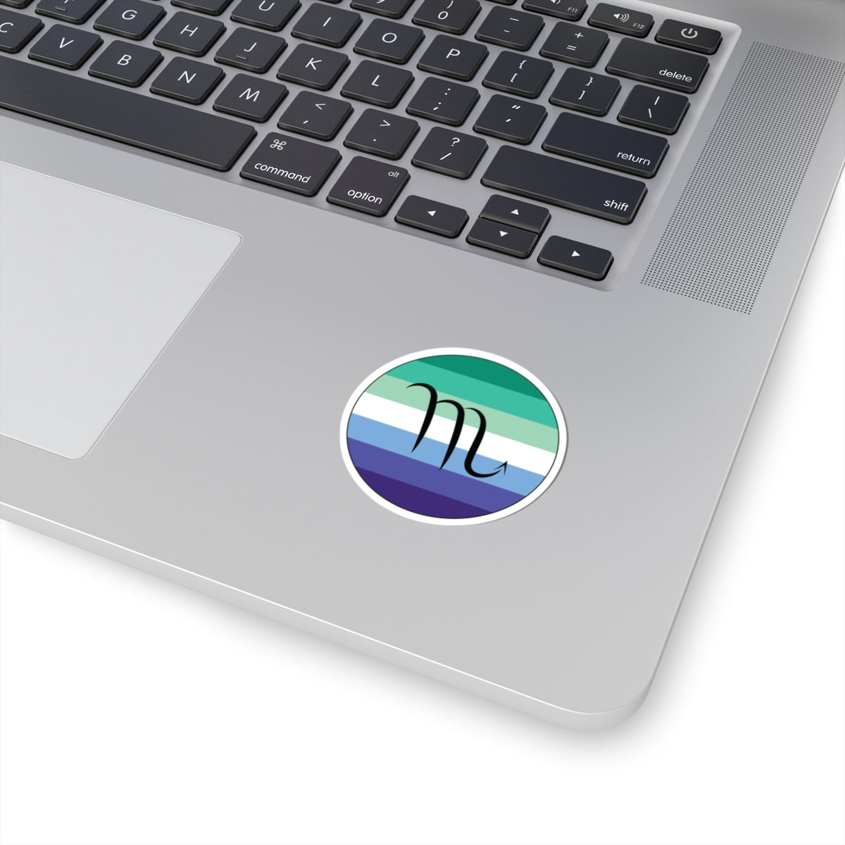 Close-up of the bottom right corner of a silver laptop featuring a 2 inch by 2 inch circular zodiac sticker with a centered Scorpio glyph on a Gay Men pride flag style background applied to the surface.