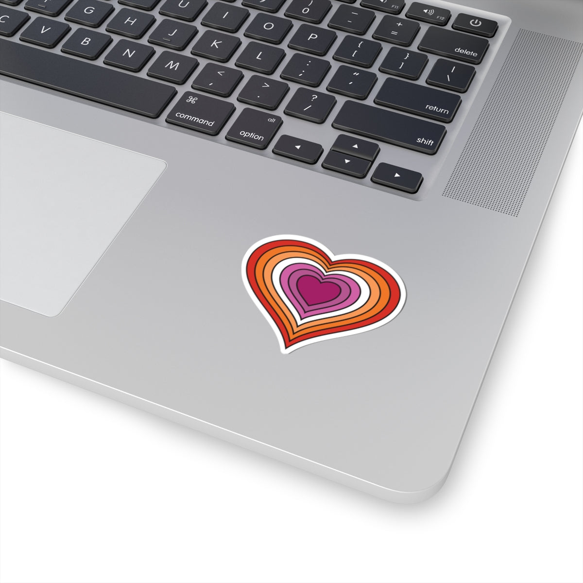 Close-up of the bottom right corner of a silver laptop featuring a 2 inches by 2 inches pride flag heart sticker in a Lesbian pride flag style applied to the surface.