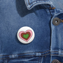 Close-up of a white circular pride button featuring a nested heart design inspired by the Gynesexual pride flag in pink, brown and green, attached to a blue denim jacket at the right chest with the button clearly visible.