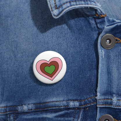 Close-up of a white circular pride button featuring a nested heart design inspired by the Gynesexual pride flag in pink, brown and green, attached to a blue denim jacket at the right chest with the button clearly visible.