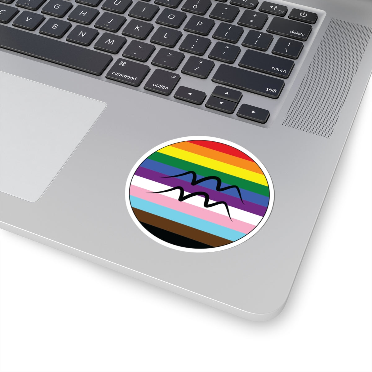 Close-up of the bottom right corner of a silver laptop featuring a 3 inch by 3 inch circular zodiac sticker with a centered Aquarius glyph on a progress pride flag style background applied to the surface.