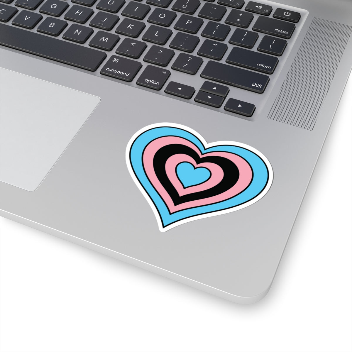 Close-up of the bottom right corner of a silver laptop featuring a 3 inch by 3 inch pride flag heart sticker in a Black Trans flag style applied to the surface.