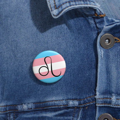 Close-up of a white circular pride button featuring the Leo zodiac glyph centered over a transgender pride flag background in light blue, pink, and white, attached to a blue denim jacket at the right chest with the button clearly visible.