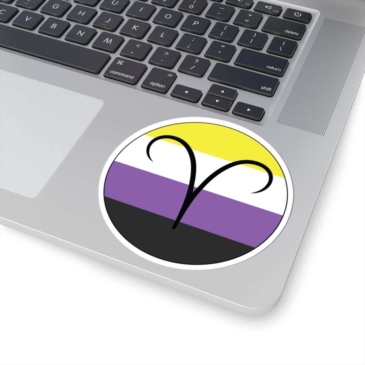 Close-up of the bottom right corner of a silver laptop featuring a 4 inch by 4 inch circular zodiac sticker with a centered Aires glyph on a Nonbinary pride flag style background applied to the surface.