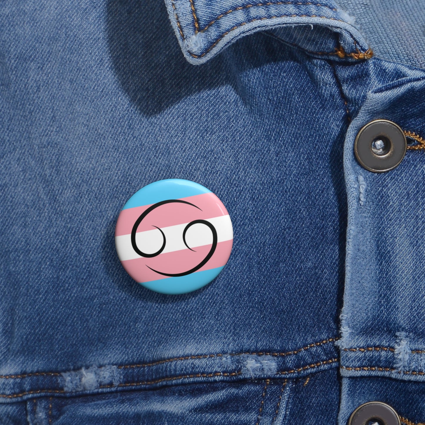 Close-up of a white circular pride button featuring the Cancer zodiac glyph centered over a transgender pride flag background in light blue, pink, and white, attached to a blue denim jacket at the right chest with the button clearly visible.