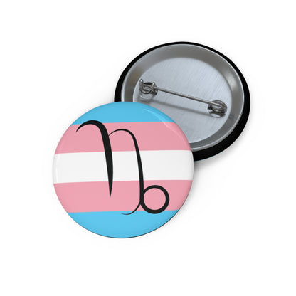Front and back view of a circular transgender pride flag button featuring the Capricorn zodiac symbol in black centered on the front.