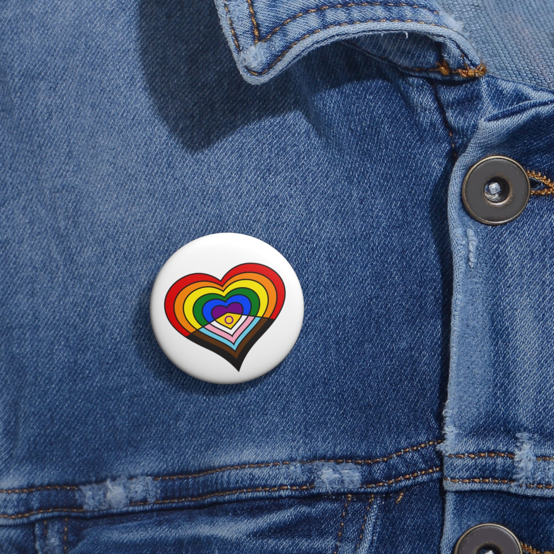 Close-up of a white circular pride button featuring a nested heart design inspired by the Intersex-Inclusive Progress pride flag in rainbow, black, brown, trans and intersex flag colors, attached to a blue denim jacket at the right chest with the button clearly visible.