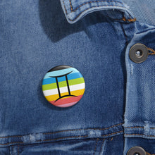 Close-up of a white circular pride button featuring the Gemini zodiac glyph centered over a queer pride flag background in pinks, yellow, white, green, blues, and black, attached to a blue denim jacket at the right chest with the button clearly visible.