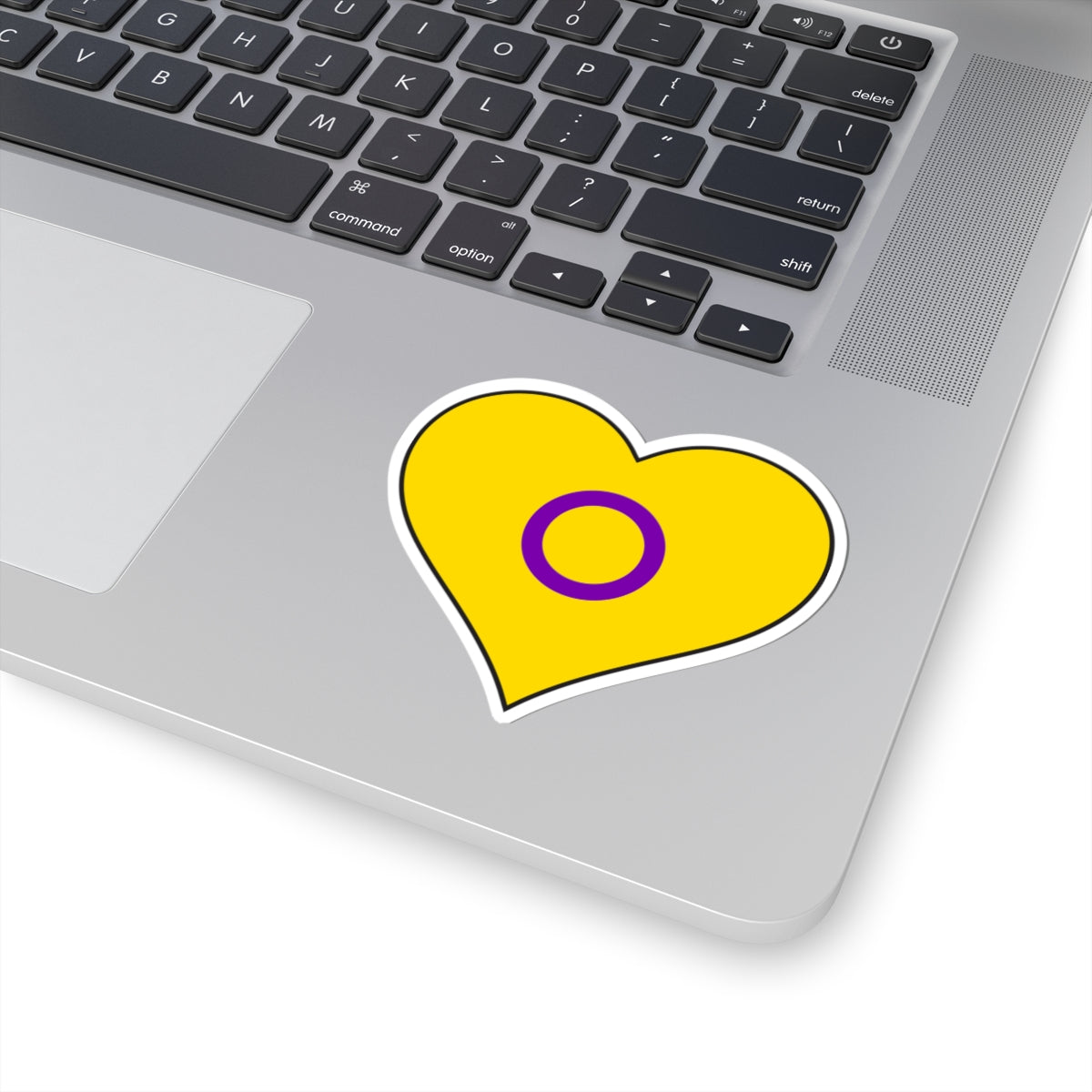 Close-up of the bottom right corner of a silver laptop featuring a 3 inch by 3 inch pride flag heart sticker in an Intersex flag style applied to the surface.