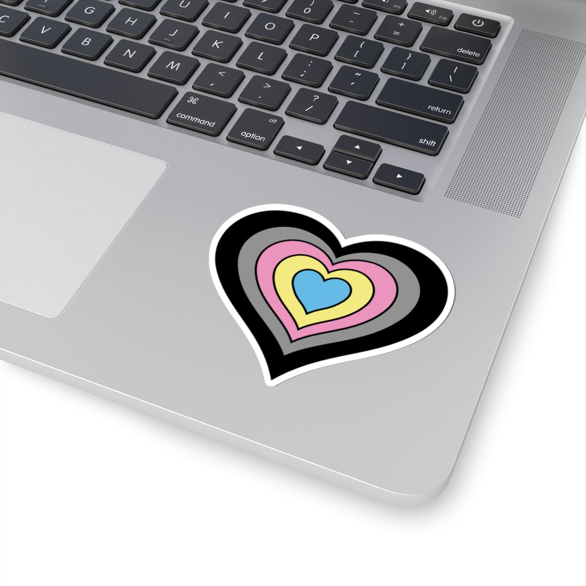 Close-up of the bottom right corner of a silver laptop featuring a 3 inch by 3 inch pride flag heart sticker in a Polygender flag style applied to the surface.