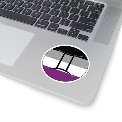 Close-up of the bottom right corner of a silver laptop featuring a 3 inch by 3 inch circular zodiac sticker with a centered Gemini glyph on an Asexual pride flag style background applied to the surface.