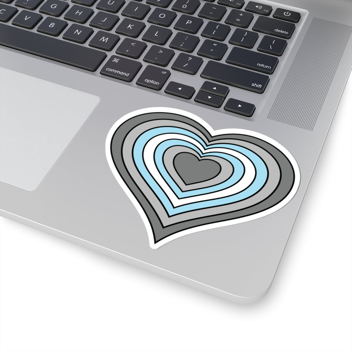 Close-up of the bottom right corner of a silver laptop featuring a 4 inch by 4 inch pride flag heart sticker in a Demiboy flag style applied to the surface.