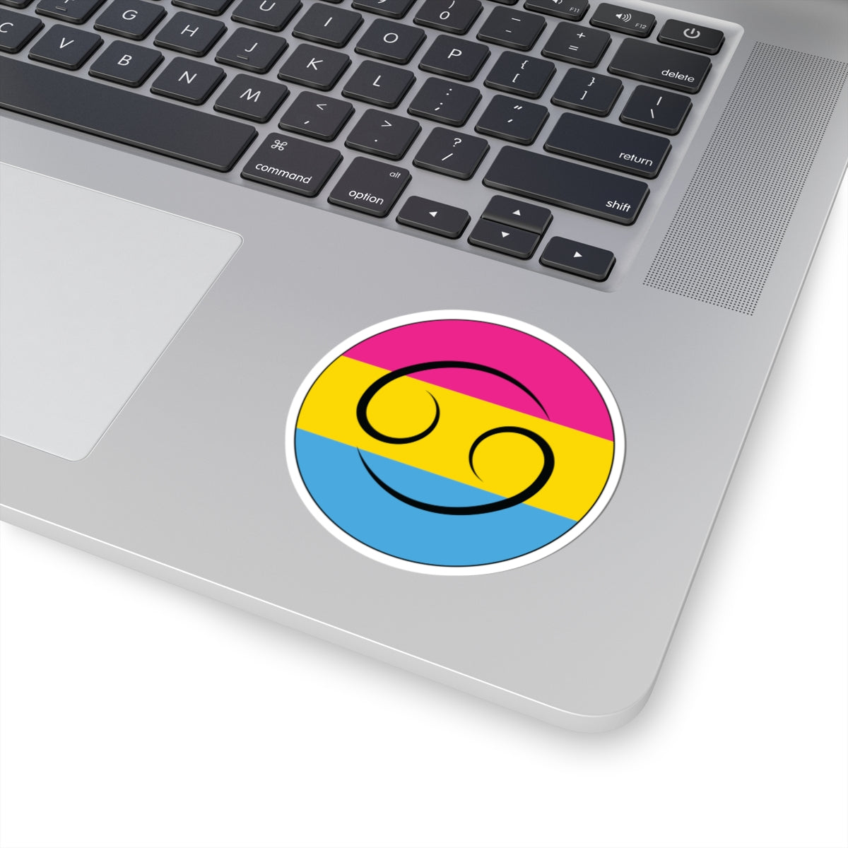 Close-up of the bottom right corner of a silver laptop featuring a 3 inch by 3 inch circular zodiac sticker with a centered Cancer glyph on a Pansexual pride flag style background applied to the surface.