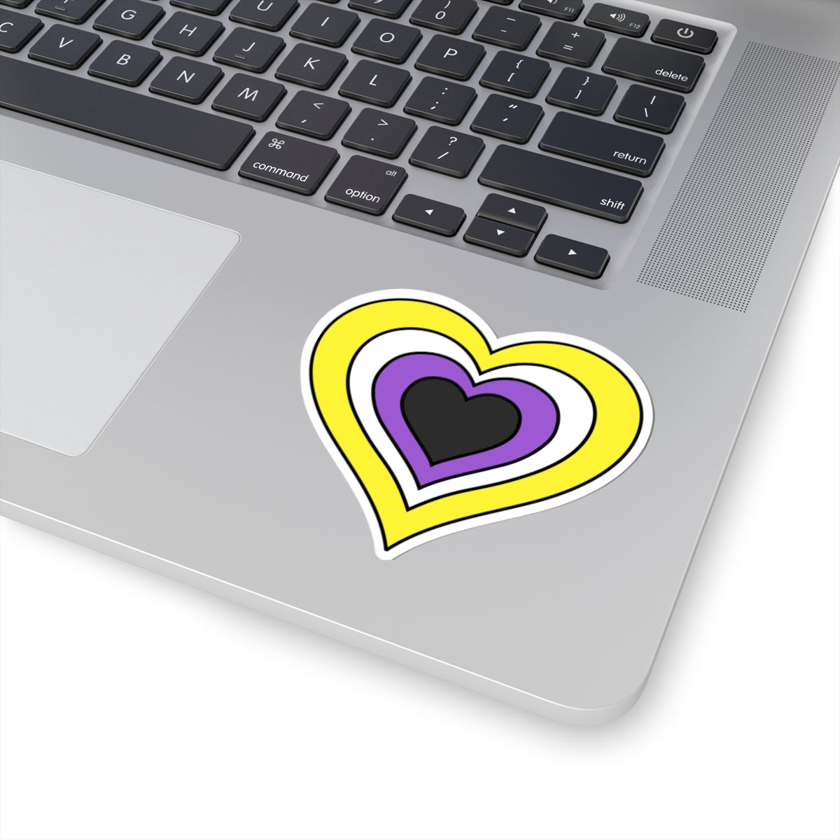 Close-up of the bottom right corner of a silver laptop featuring a 3 inch by 3 inch pride flag heart sticker in a Nonbinary flag style applied to the surface.