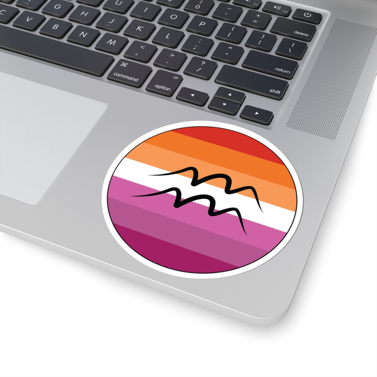 Close-up of the bottom right corner of a silver laptop featuring a 4 inch by 4 inch circular zodiac sticker with a centered Aquarius glyph on a Lesbian pride flag style background applied to the surface.