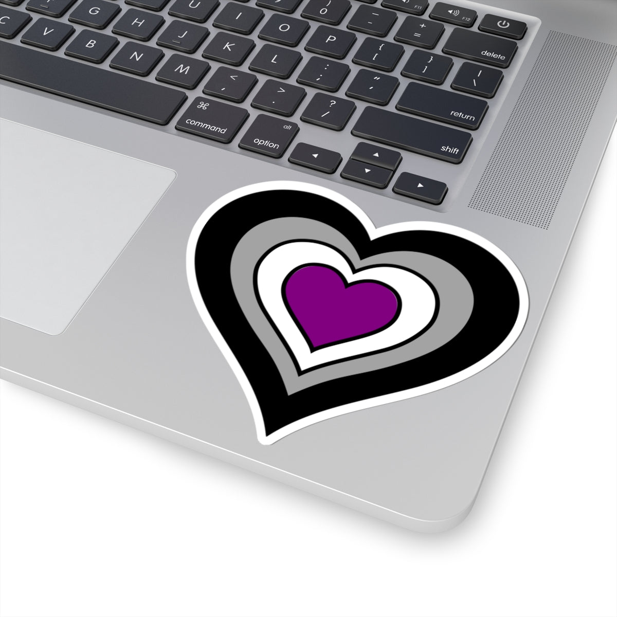 Close-up of the bottom right corner of a silver laptop featuring a 4 inch by 4 inch pride flag heart sticker in an Asexual flag style applied to the surface.