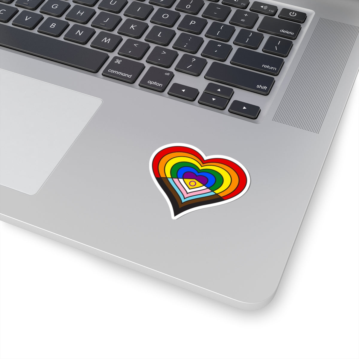 Close-up of the bottom right corner of a silver laptop featuring a 2 inch by 2 inch pride flag heart sticker in an Intersex-Inclusive Progress pride flag style applied to the surface.