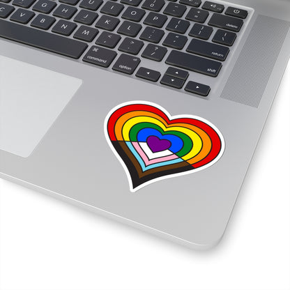 Close-up of the bottom right corner of a silver laptop featuring a 3 inch by 3 inch pride flag heart sticker in a Progress pride flag style applied to the surface.