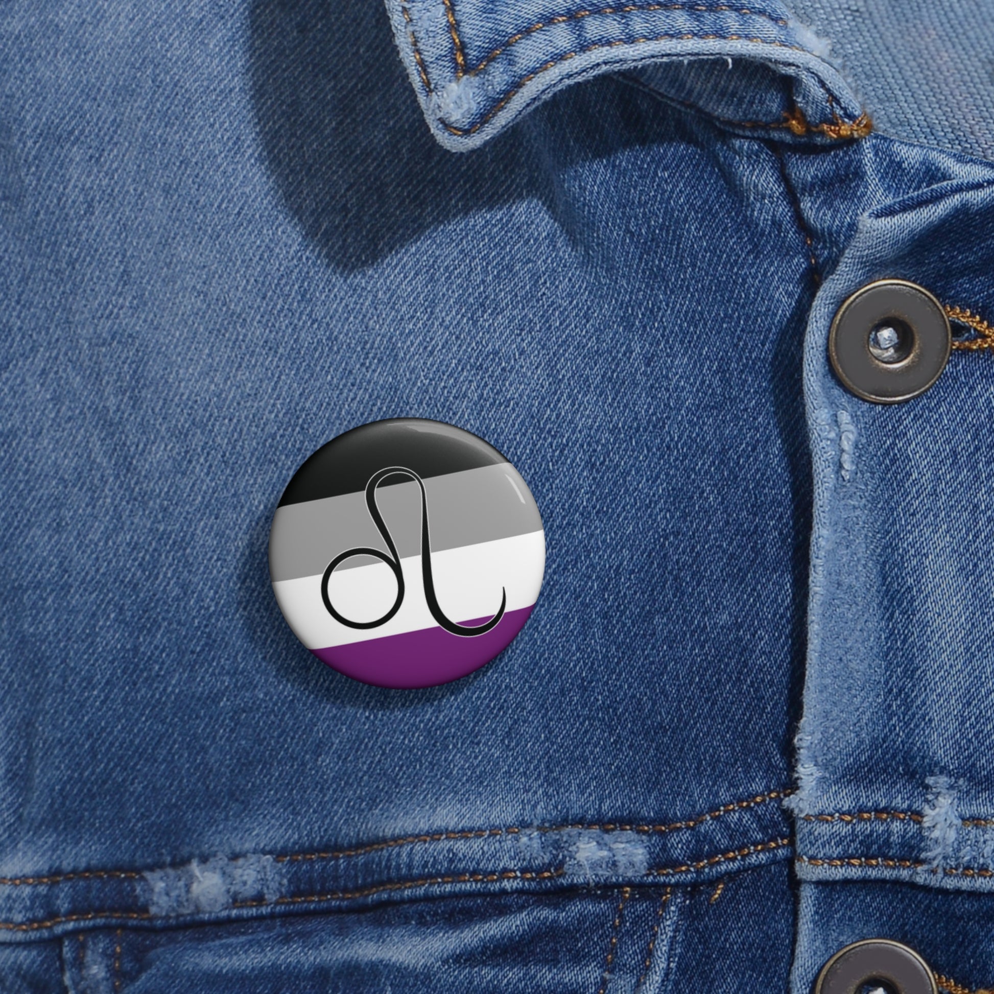 Close-up of a white circular pride button featuring the Leo zodiac glyph centered over an asexual pride flag background in black, gray, white, and purple, attached to a blue denim jacket at the right chest with the button clearly visible.