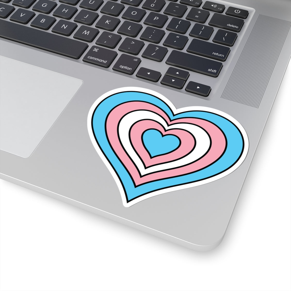 Close-up of the bottom right corner of a silver laptop featuring a 4 inch by 4 inch pride flag heart sticker in a Transgender flag style applied to the surface.