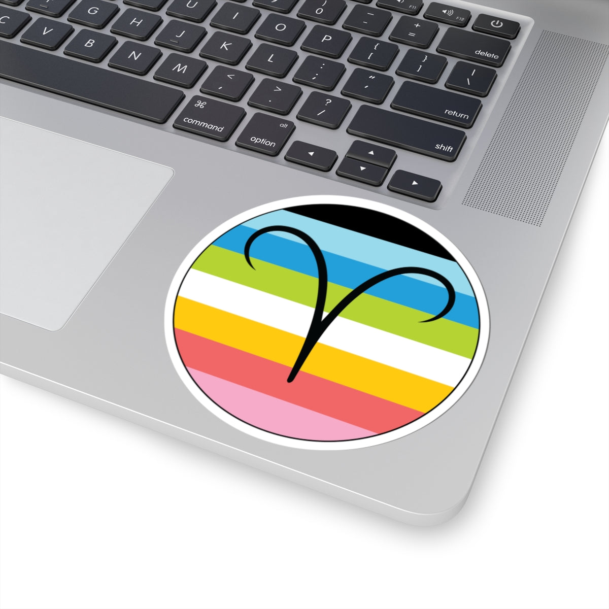 Close-up of the bottom right corner of a silver laptop featuring a 4 inch by 4 inch circular zodiac sticker with a centered Aires glyph on a Queer pride flag style background applied to the surface.